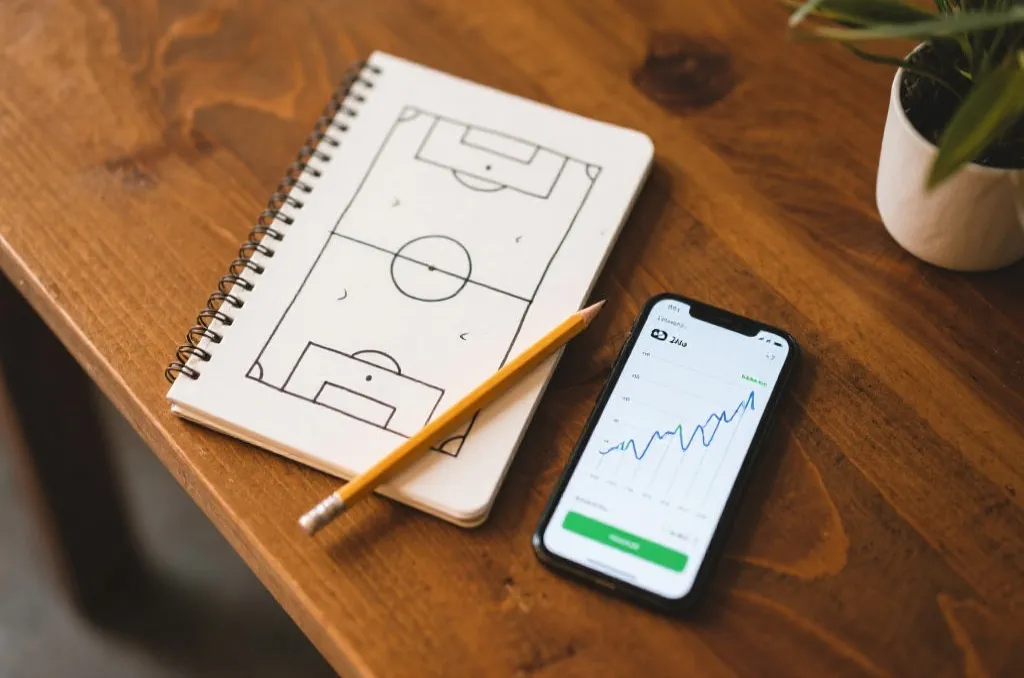A tactical notebook with hand-drawn football pitch diagrams, a pencil, and a smartphone displaying a clean statistical line graph on a wooden table